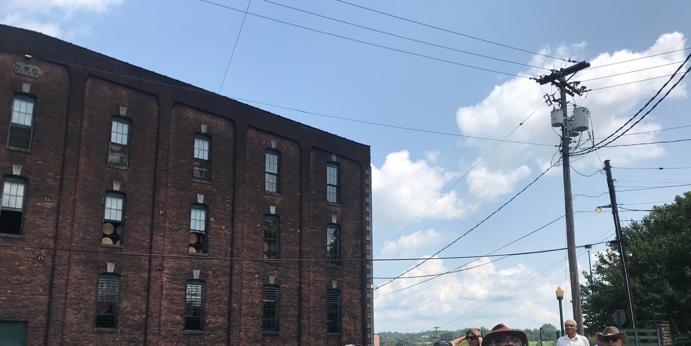 A long brick barrel-ageing warehouse at Buffalo Trace Distillery in Frankfort, Kentucky, photographed from the roadside on a bright day, its regular bays of small windows running the length of the building
