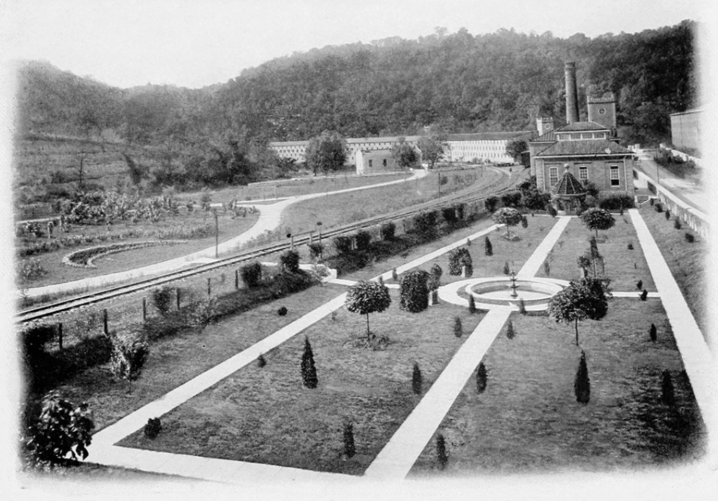 A 1916 photograph of the Old Taylor distillery in Millville, Kentucky: formal sunken gardens with clipped hedges and gravel paths in the foreground, a stone springhouse at right, long warehouse buildings and a tall brick stack in the valley behind, wooded hills beyond